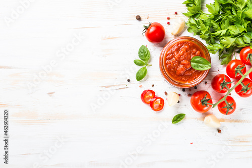 Traditional italian tomato sauce in glass jar with herbs and spices at white kitchen table. Top view with copy space.