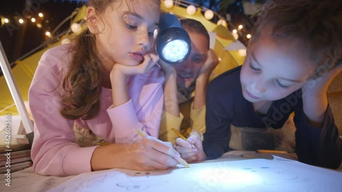 Brother and sister draw while daddy holds glowing flashlight spending time together by play tent with fairy lights in cottage yard in evening closeup photo