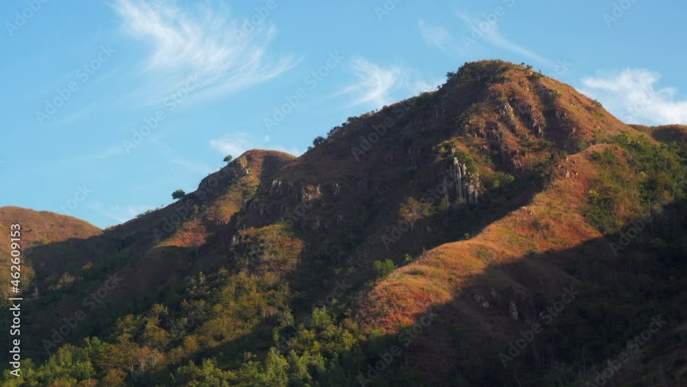 Old brown mountains with green trees growing on slopes stand against blue sky with floating light white clouds on nice sunny day Old brown mountains with green trees growing on slopes stand against blue sky with floating light white clouds on nice sunny day