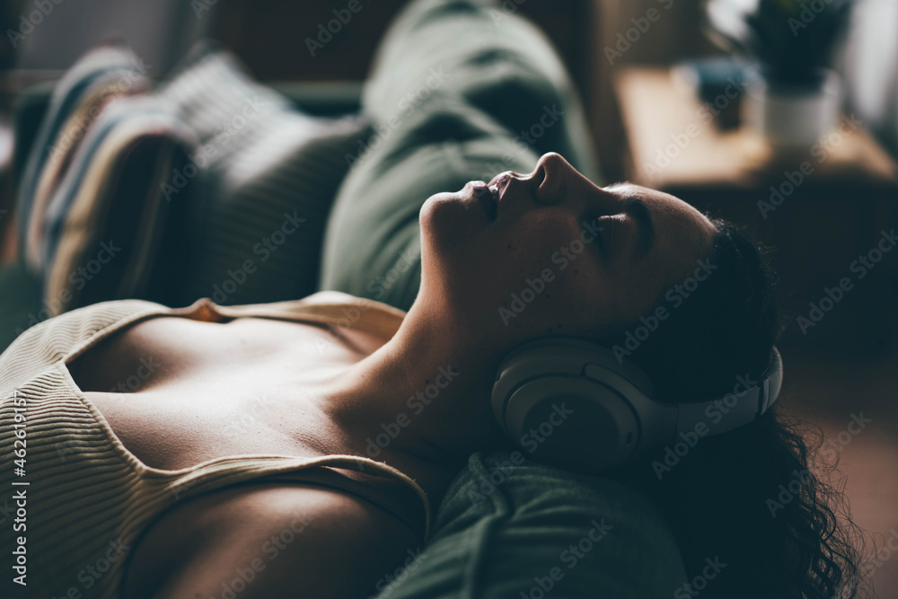 Curly haired overweight young woman wearing comfortable clothes lies on green sofa listening to music in wireless headphones in stylish room - obrazy, fototapety, plakaty 