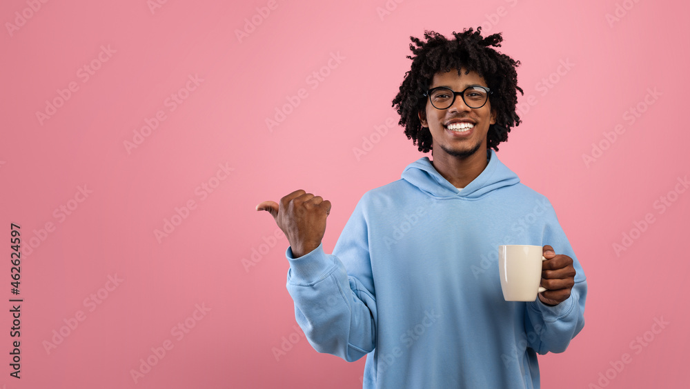 Handsome Afro teen guy in casual wear pointing aside at empty space, holding cup of coffee on pink background Handsome Afro teen guy in casual wear pointing aside at empty space, holding cup of coffee on pink background