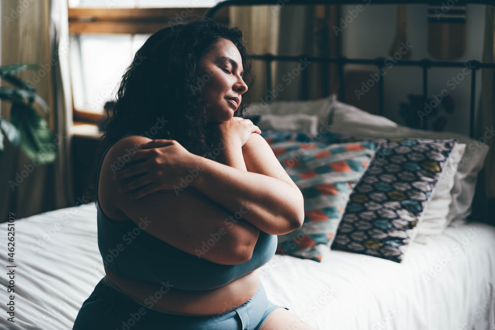Obraz na plátně Curly haired overweight young woman in blue top and shorts with satisfaction on face accepts curvy body shape in stylish bedroom
