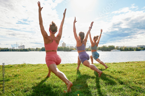 Three fit young beautiful women doing Reverse Warrior Pose  Crescent lunge variation  Viparita Virabhadrasana  working out in park on summer day  wearing sportswear tops  full length. Fitness  sport