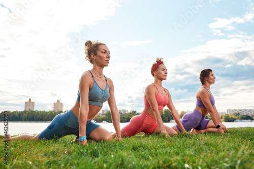 Three fit young beautiful women doing Reverse Warrior Pose, Crescent lunge variation, Viparita Virabhadrasana, working out in park on summer day, wearing sportswear tops, full length. Fitness, sport