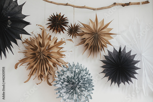 Stylish Christmas stars hanging from wooden branch on white wall. Modern festive scandinavian decor in room. Simple swedish paper stars. Winter holiday preparations. Merry Christmas!