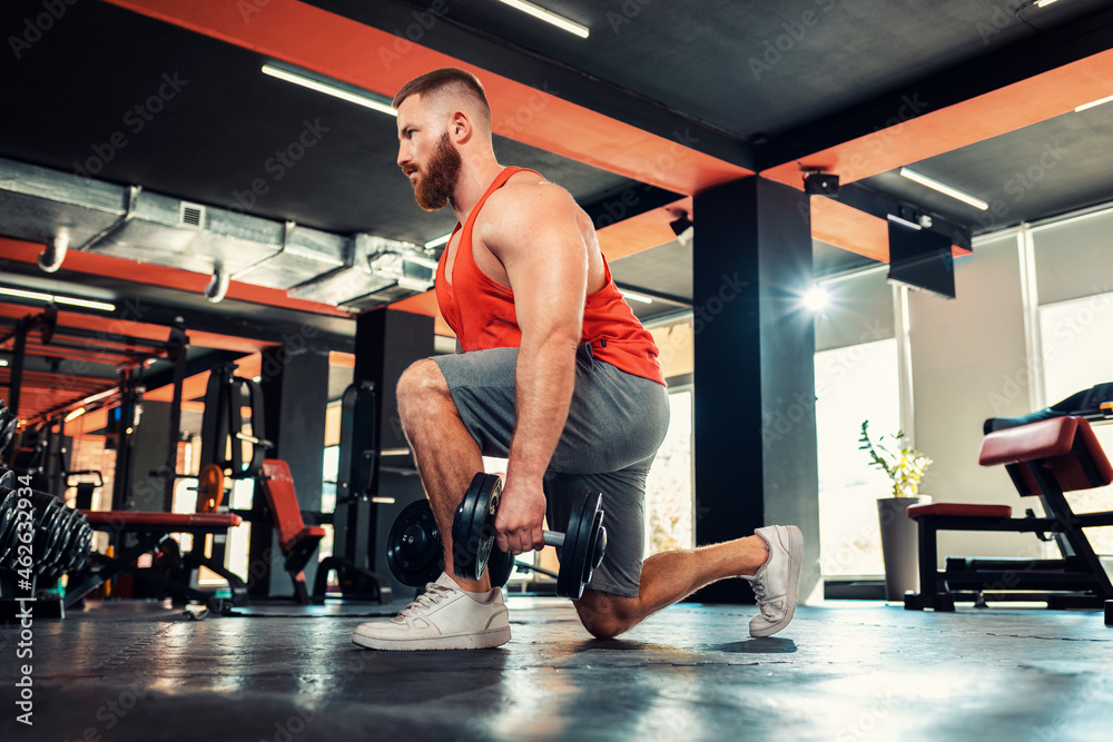 A young bearded athletic man trains with dumbbells in the gym. Squat exercise. Bottom view. The concept of fitness and workout A young bearded athletic man trains with dumbbells in the gym. Squat exercise. Bottom view. The concept of fitness and workout