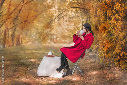 A girl in a red coat with a book drinks tea in the autumn forest