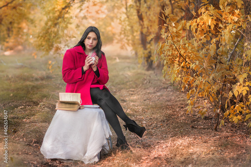 A girl in a red coat with a book drinks tea in the autumn forest
