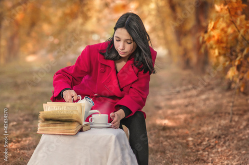 A girl in a red coat with a book drinks tea in the autumn forest