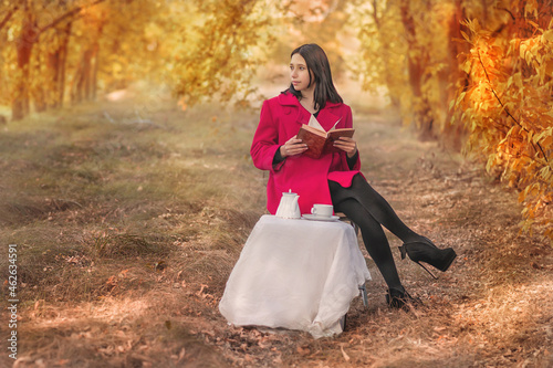A girl in a red coat with a book drinks tea in the autumn forest