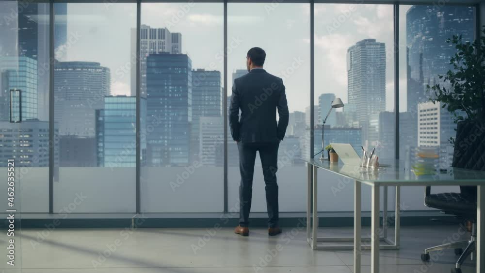 Confident Young Businessman in a Perfect Tailored Suit Standing in His Modern Office Looking out of the Window on Big City with Skyscrapers. Successful Finance Manager Planning Project Strategy. Confident Young Businessman in a Perfect Tailored Suit Standing in His Modern Office Looking out of the Window on Big City with Skyscrapers. Successful Finance Manager Planning Project Strategy.