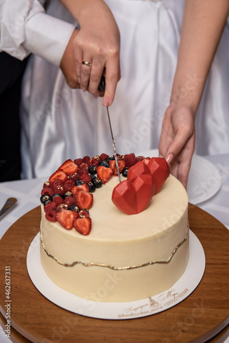 Newlyweds are cutting a beautiful white cake with berries and heart-shaped decor. Close-up
