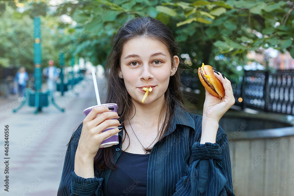 young brunette holds a hamburger and a glass with a drink in her hands. French fries are clamped in her teeth