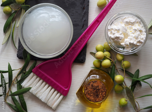 Ingredients and homemade hair mask. A bowl with a mask, a brush for applying a mask. Olive branch with leaves and olives. White background. Flat lay.