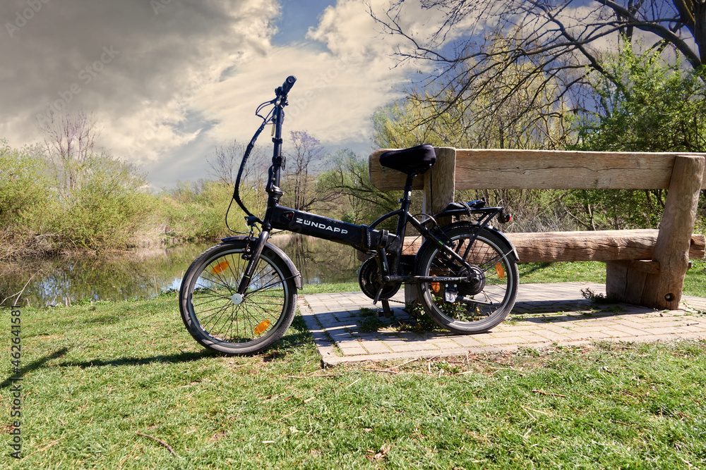 Zündapp electric folding bike leaning against a massive rustic wooden bench  in front of a river under sensible cloudy sky in Gifhorn, Germany, April  18, 2020 Stock Photo | Adobe Stock