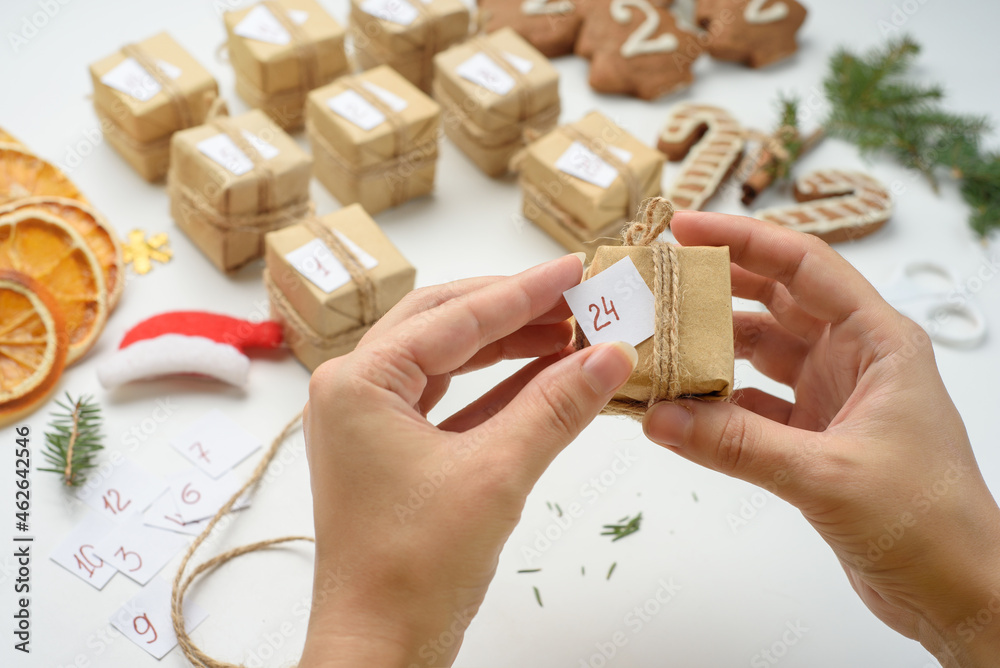 Fototapeta premium Female hands packages gifts for advent calendar. Preparing for Christmas with gingerbread cookies, dried oranges, sprigs of fir. Eco friendly new year holidays