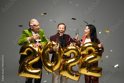 Excited interracial friends with champagne standing near balloons in shape of 2022 numbers and confetti on grey background