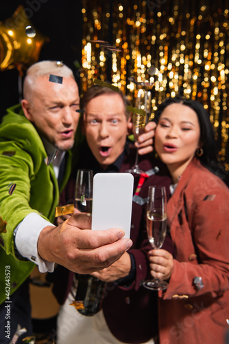 Smartphone in hand of blurred man taking selfie with interracial friends and champagne during new year party on black background
