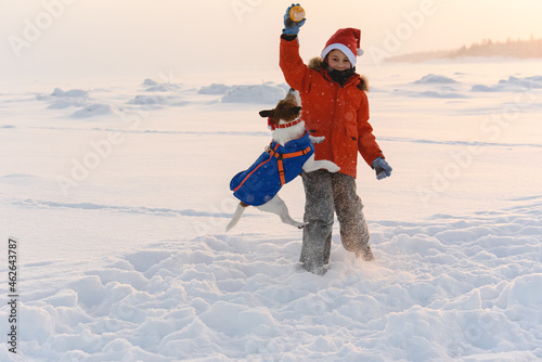 Boy wearing warm clothing and Santa Claus hat playing with his pet dog on a beach of White sea on December day photo