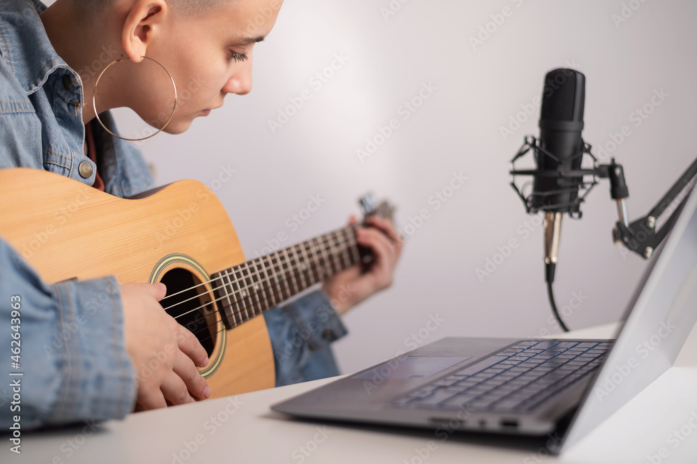 Fototapeta premium Young caucasian woman with short blonde hair playing guitar and streaming live on laptop.
