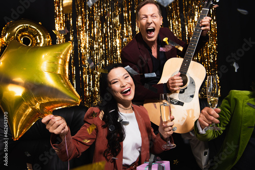 Cheerful asian woman holding balloon and champagne near friends with acoustic guitar and confetti on black background