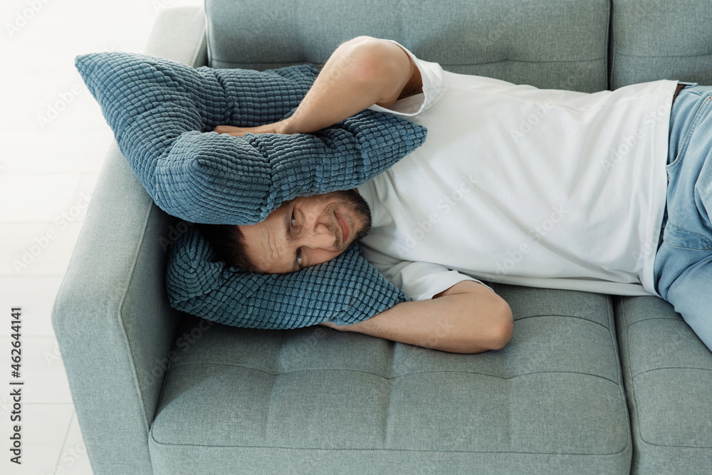 Unhappy man cover ears with pillows to avoid noise from neighbor while napping on couch. Frustrated guy lying on sofa annoyed with loud noisy sounds in apartment. Dissatisfied male trying to sleep Unhappy man cover ears with pillows to avoid noise from neighbor while napping on couch. Frustrated guy lying on sofa annoyed with loud noisy sounds in apartment. Dissatisfied male trying to sleep