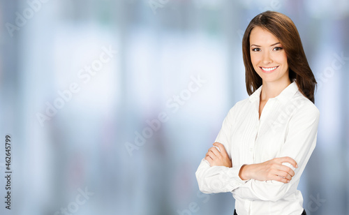 Portrait of smiling businesswoman in crossed arms pose, confident white clothing. Business concept. Brunette business woman, indoors. Blurred modern office interior background.
