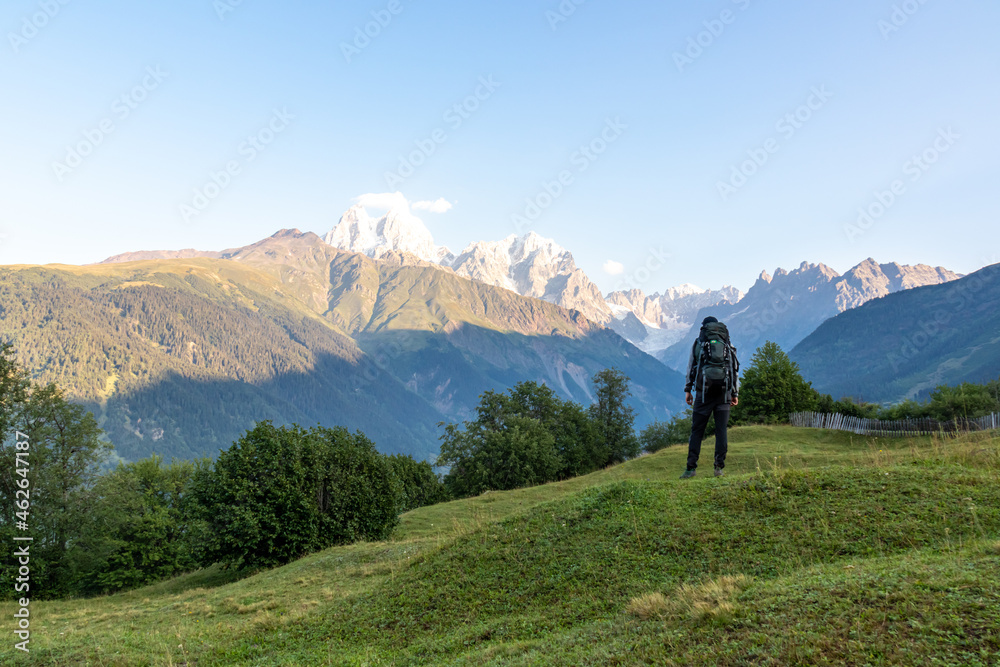 A man with a big hiking backpack enjoying the view on the first sunbeams reaching the peaks of Ushba in Caucasus, Georgia. Cloudless sky above the high, snow-capped mountains. Green meadow.