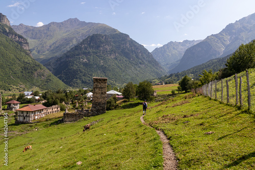 A narrow pathway leading to Zhabeshi, a mountain village in Georgia. A few lookout towers towering above the village. High Caucasus mountain chains. lush green pastures with a few cows grazing on them photo
