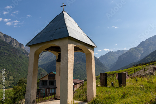 A small bell tower on a lush green pasture in Caucasus, Georgia. There are high mountain chains around. Clear and bright day. Spirituality and serenity. photo