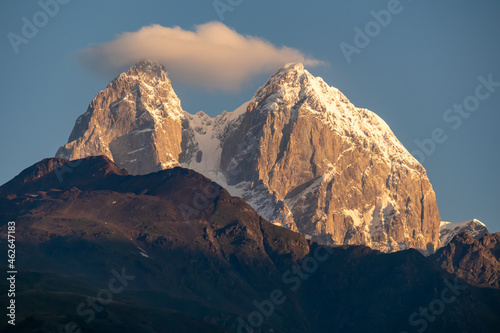 First sunrise reaching the peaks of Ushba in Caucasus, Georgia. Cloudless sky above the high and snow-capped mountains. The hills below are shrouded with shadows. Daybreak. High and desolate mountains