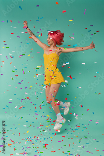 full length of cheerful young woman with pink dyed hair jumping near colorful confetti on blue