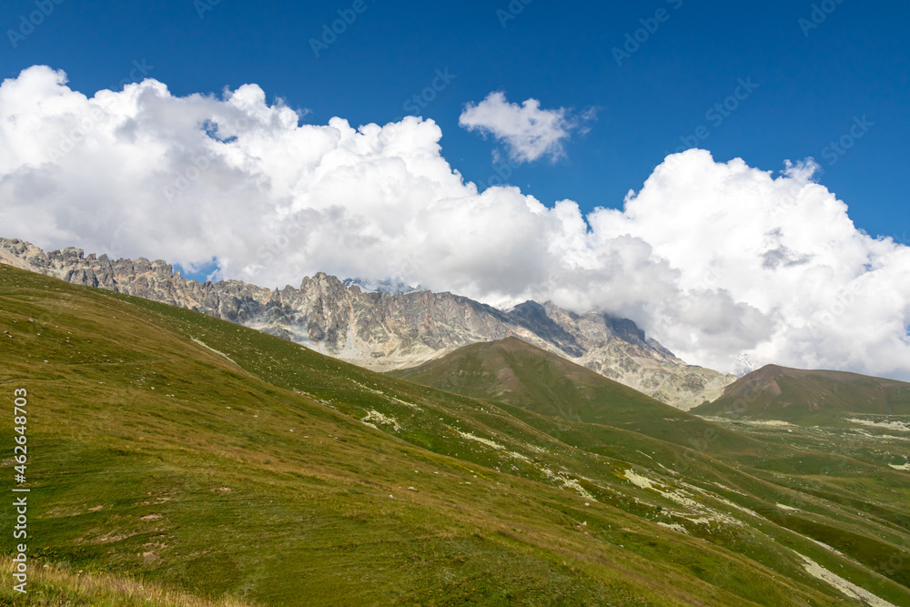 A panoramic view on high Caucasus mountains in Georgia. There are high glaciers in the back. Thick clouds above the sharp peaks. Lush pastures on the sides. Barren peaks.  Idyllic landscape.