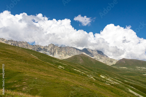 A panoramic view on high Caucasus mountains in Georgia. There are high glaciers in the back. Thick clouds above the sharp peaks. Lush pastures on the sides. Barren peaks. Idyllic landscape.