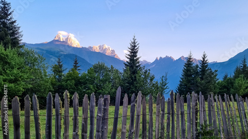 A wooden fence in front of a lush pasture with a view on the first sunbeams reaching the peaks of Ushba in Caucasus, Georgia. Cloudless sky above the high and snow-capped mountains. Daybreak.