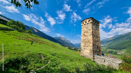 A man walking next to a lookout tower on a lush green pasture in Caucasus, Georgia. There are high mountain chains around. Clear and bright day with few clouds. Defense building. Hiking remedy photo