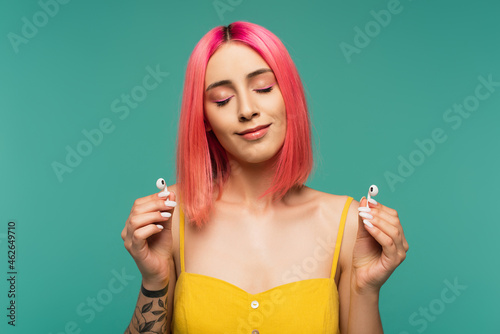 pleased young woman with pink dyed hair holding wireless earphones isolated on blue