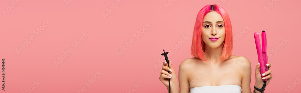 happy young woman with colorful hair holding hair straightener isolated on pink, banner