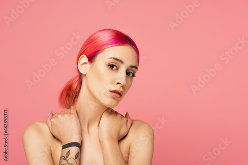 tattooed young woman with colorful hair touching bare shoulders while looking at camera isolated on pink