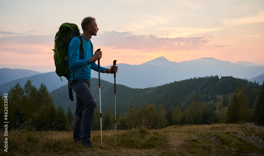 Man traveler against backdrop of mountain hills and pink cloudy evening sky at sunset. Hiker with trekking poles and touristic backpack standing on glade and admiring mountain fairytale landscape.
