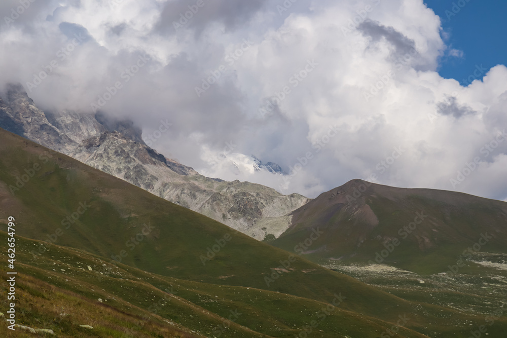A panoramic view on high Caucasus mountains in Georgia. There are high, snowcapped peaks in the back. Lush pasture in front. Idyllic landscape. Thic clouds above the sharp peaks. Natural remedy