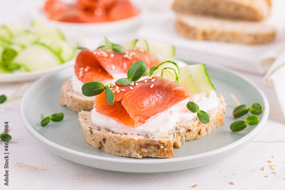Toasts or open sandwiches with salted salmon, ricotta cream cheese, cucumber, sprouts and whole grain bread on white table. Healthy food, diet. Breakfast.