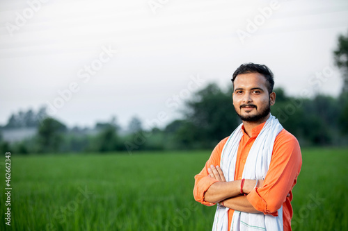 Happy Indian young man farmer giving side pose standing in field with hands crossed wearing kurta  smiling village male peasant looking on camera in farm  greenery  blur background  copy space.