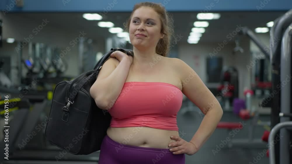 Portrait of smiling confident plus-size woman walking in gym looking around smiling. Front view of happy overweight Caucasian young sportswoman with bag strolling examining sports equipment Portrait of smiling confident plus-size woman walking in gym looking around smiling. Front view of happy overweight Caucasian young sportswoman with bag strolling examining sports equipment