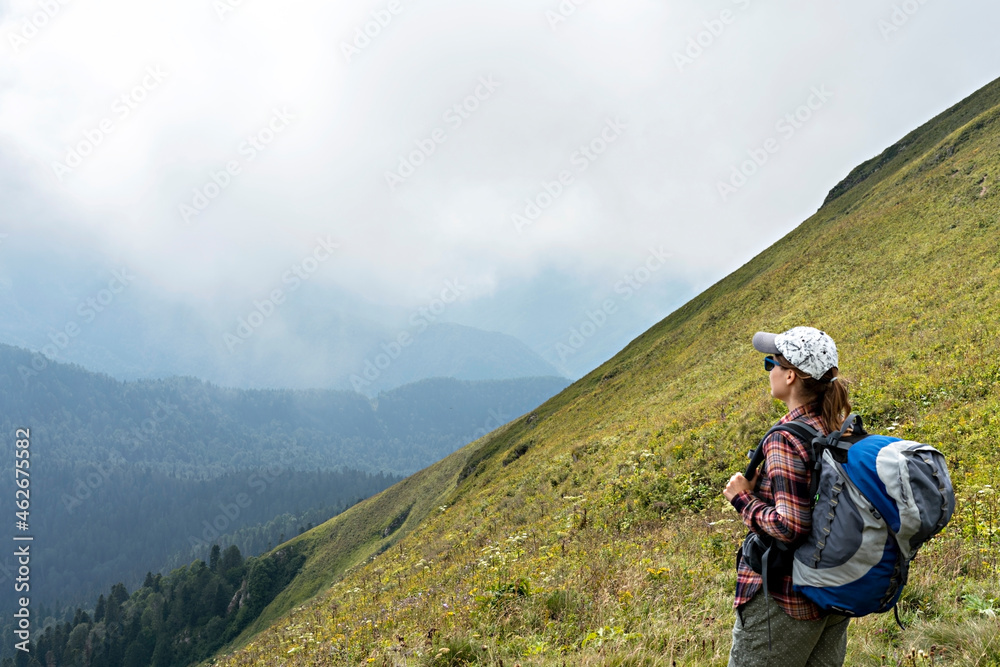 Young woman hiker in cap and sunglasses with large hiking backpack looking at the mountain view of the Aibga ridge of Caucasus mountains, healthy active lifestyle, weekend activities beauty in nature