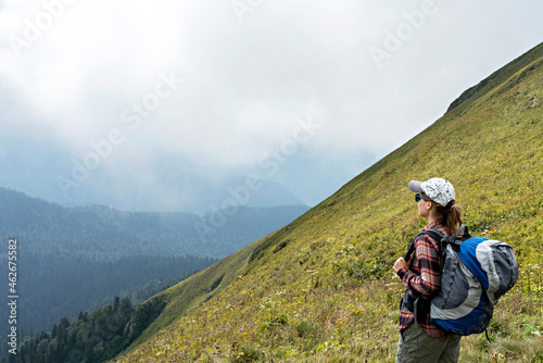 Young woman hiker in cap and sunglasses with large hiking backpack looking at the mountain view of the Aibga ridge of Caucasus mountains  healthy active lifestyle  weekend activities beauty in nature