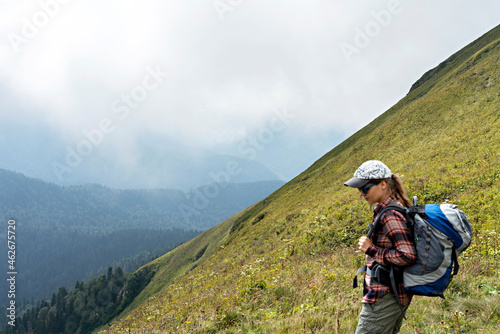 Young woman hiker in cap and sunglasses with large hiking backpack looking at mountain view of the Aibga ridge of the Caucasus mountains, healthy active lifestyle, weekend activities beauty in nature