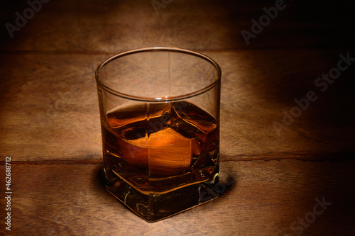 A glass of whiskey on a wooden table in a dark room lit by a weak light