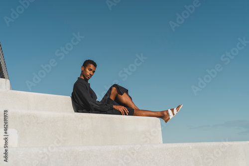 Young man wearing black kaftan sitting on concrete stairs under blue sky photo