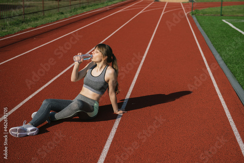 Sportswoman sitting on racetrack and drinking water after workout